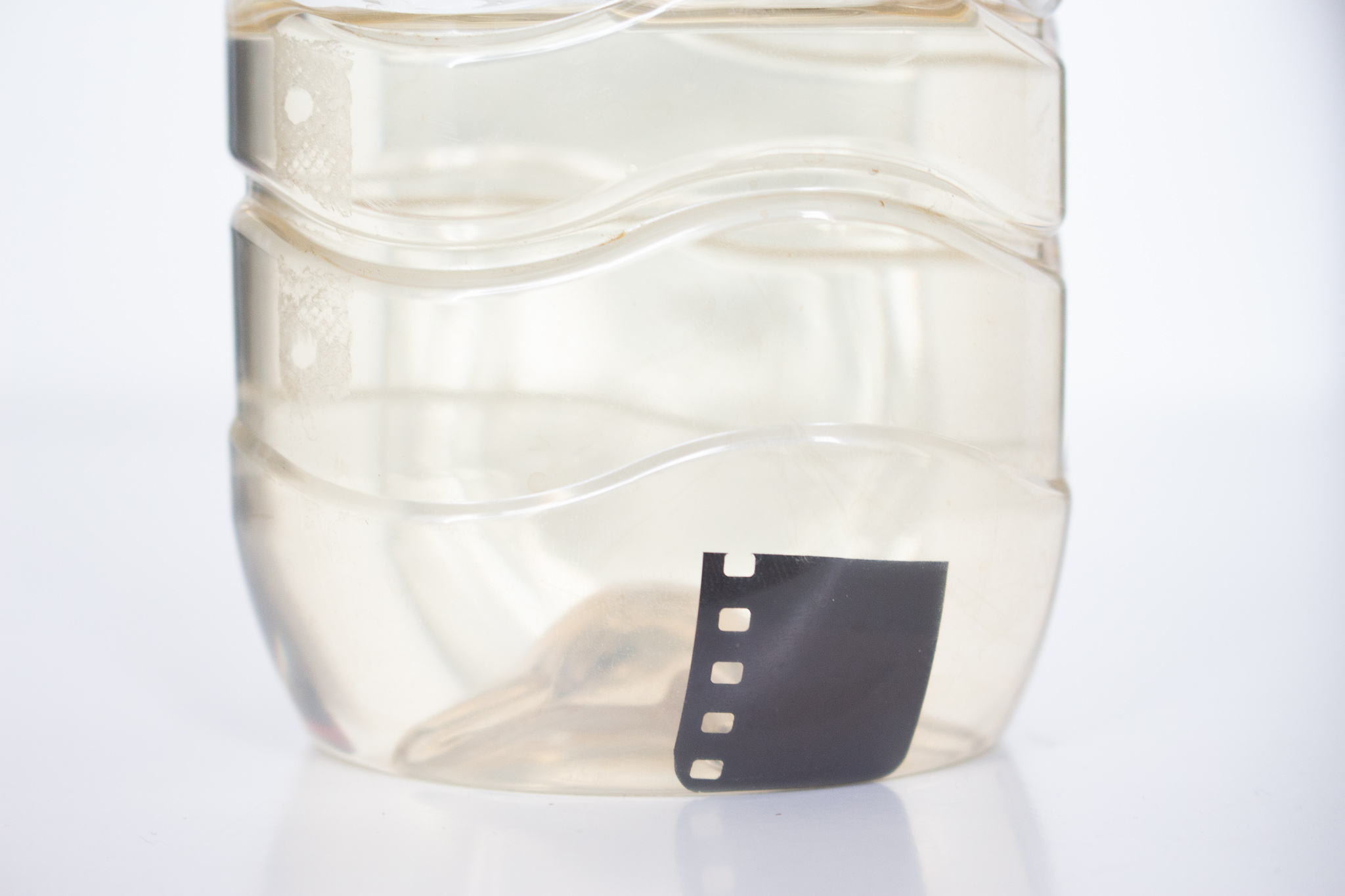 Photographie d'un morceau de pellicule noircie dans une bouteille transparente remplie de révélateur